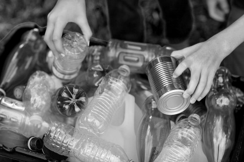 A close-up black and white image showing two hands collecting empty plastic bottles and a metal can from a pile of waste. The plastic bottles vary in size and shape, with some having ridged surfaces and others smooth, and include both clear and slightly tinted ones. The metal can being held is cylindrical with a smooth, reflective surface. The waste items are situated on a flat surface outdoors, possibly on a paved area or near a skip, consistent with rubbish removal activities. The background shows additional bottles and recyclable materials, emphasizing the focus on waste separation and collection, typical of rubbish clearance services in Fulham. The lighting is natural, creating slight shadows and highlights on the different materials, capturing the textures and details of the recyclable rubbish. Office Clearance Fulham’s rubbish removal service would handle such waste disposal tasks efficiently in the local area.