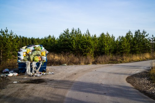 A large blue wheeled bin filled with a mixture of household waste, including plastic bags, cardboard boxes, and recyclable materials, is positioned on the gravel edge of a rural, unpaved road. The bin's lid is open, revealing the refuse inside, which appears loosely packed and spilling over in some areas. Surrounding the bin, there are scattered items and smaller pieces of waste on the ground, suggesting an accumulation of rubbish awaiting collection. In the background, there is a dense line of green trees and shrubbery, indicating a countryside setting near Fulham or west London, with clear blue skies above. The scene illustrates improper or abandoned rubbish disposal, highlighting the importance of professional waste removal services, such as those provided by Office Clearance Fulham, to maintain cleanliness and environmental standards in the area.