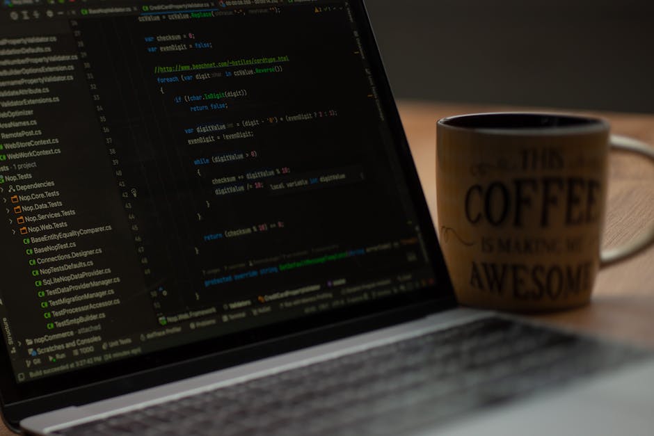 A person wearing a light blue shirt is seated at a desk, working on a laptop with a black keyboard. The laptop screen displays lines of colorful code in red, blue, yellow, and white, indicating progra